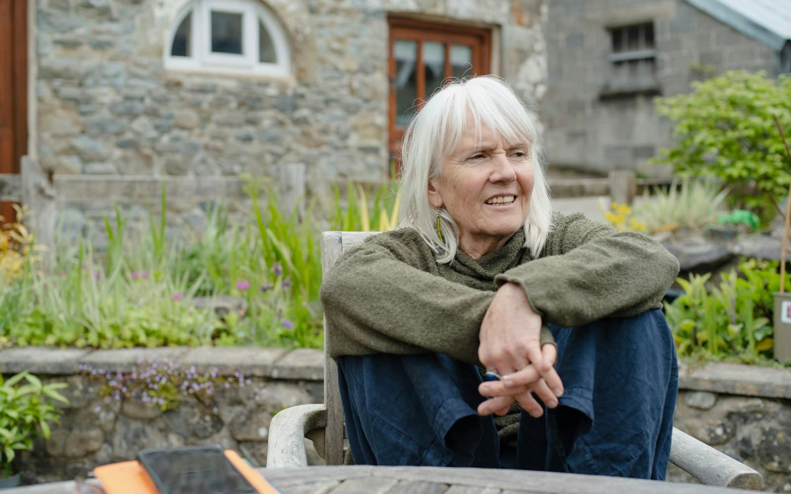 A white-haired woman smiling at the day in the garden of a stone house